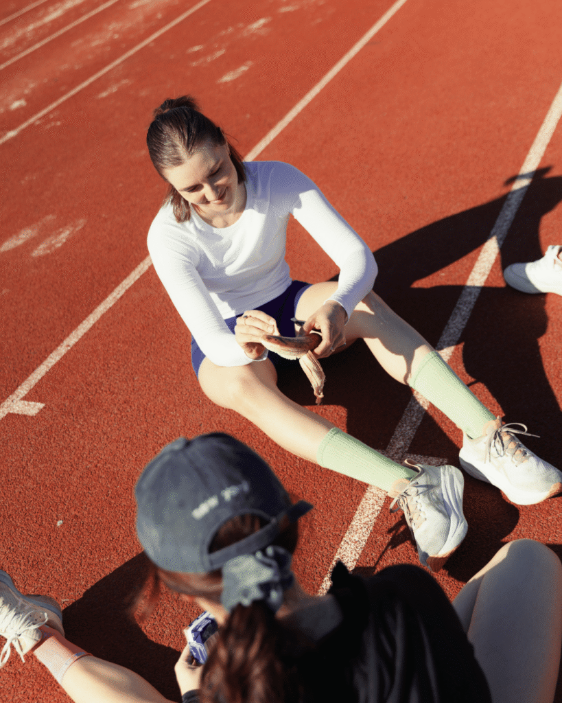 An athlete sitting on the track peeling a banana while chatting with teammates after a training session