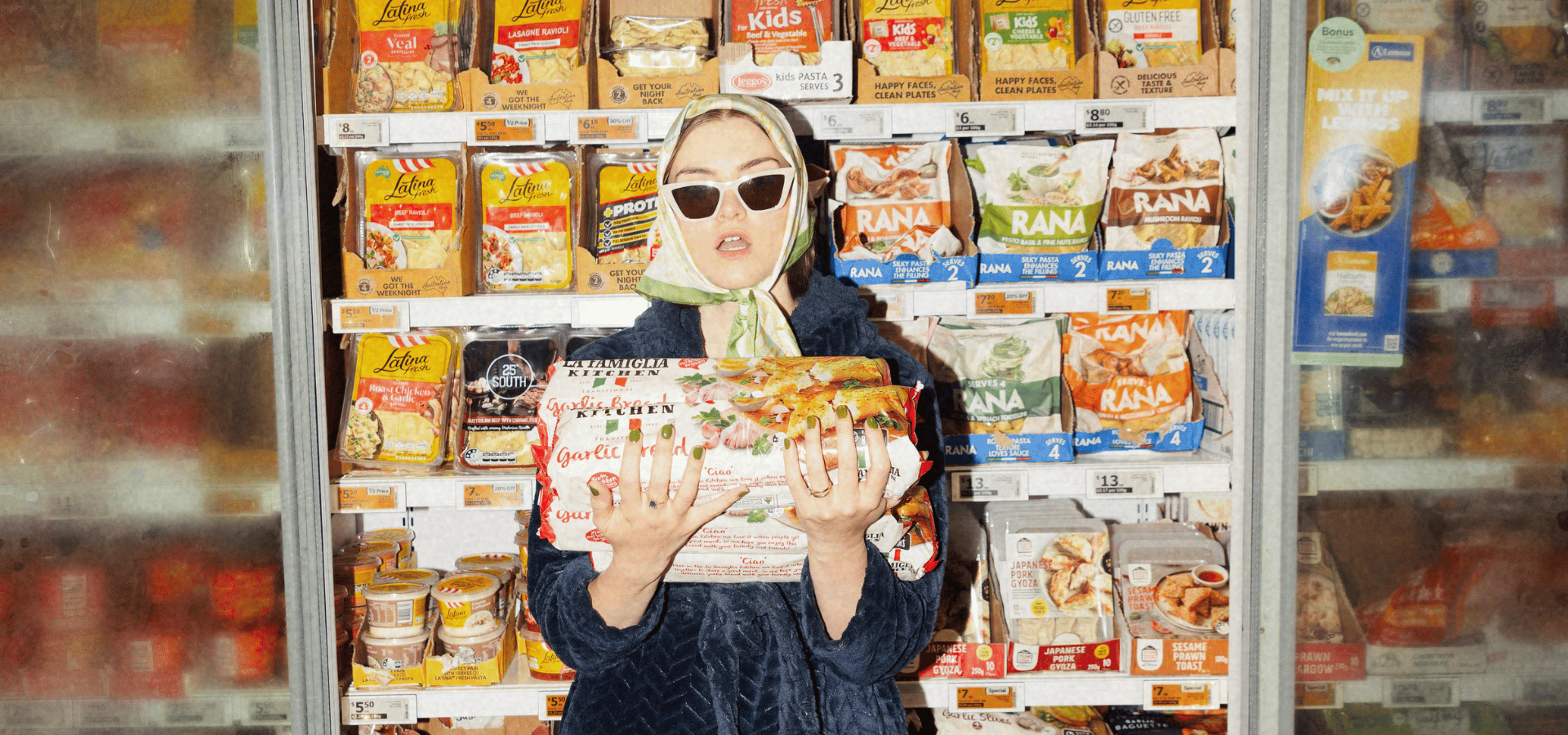Woman in sunglasses and headscarf holding packaged frozen meals in a supermarket freezer aisle
